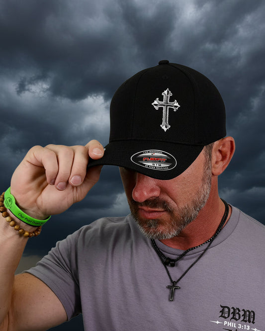A man wearing a black baseball cap with a silver embroidered cross on the front, holding the brim. He is wearing a gray t-shirt and a necklace with a cross pendant, against a plain white background.