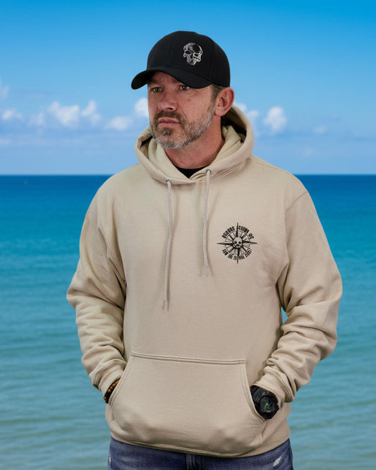 Man wearing a beige hoodie and black cap standing in front of a blue ocean.