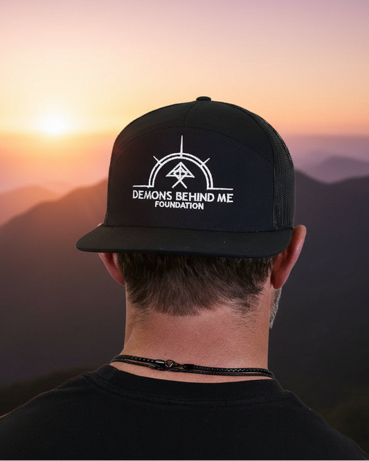 A man viewed from behind wearing a black snapback trucker hat with a white graphic logo that reads 'DEMONS BEHIND ME FOUNDATION' on the front panel.