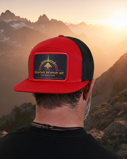 A man viewed from the back wearing a red and black trucker hat with a black patch featuring a gold emblem and the text 'DEMONS BEHIND ME FOUNDATION'.