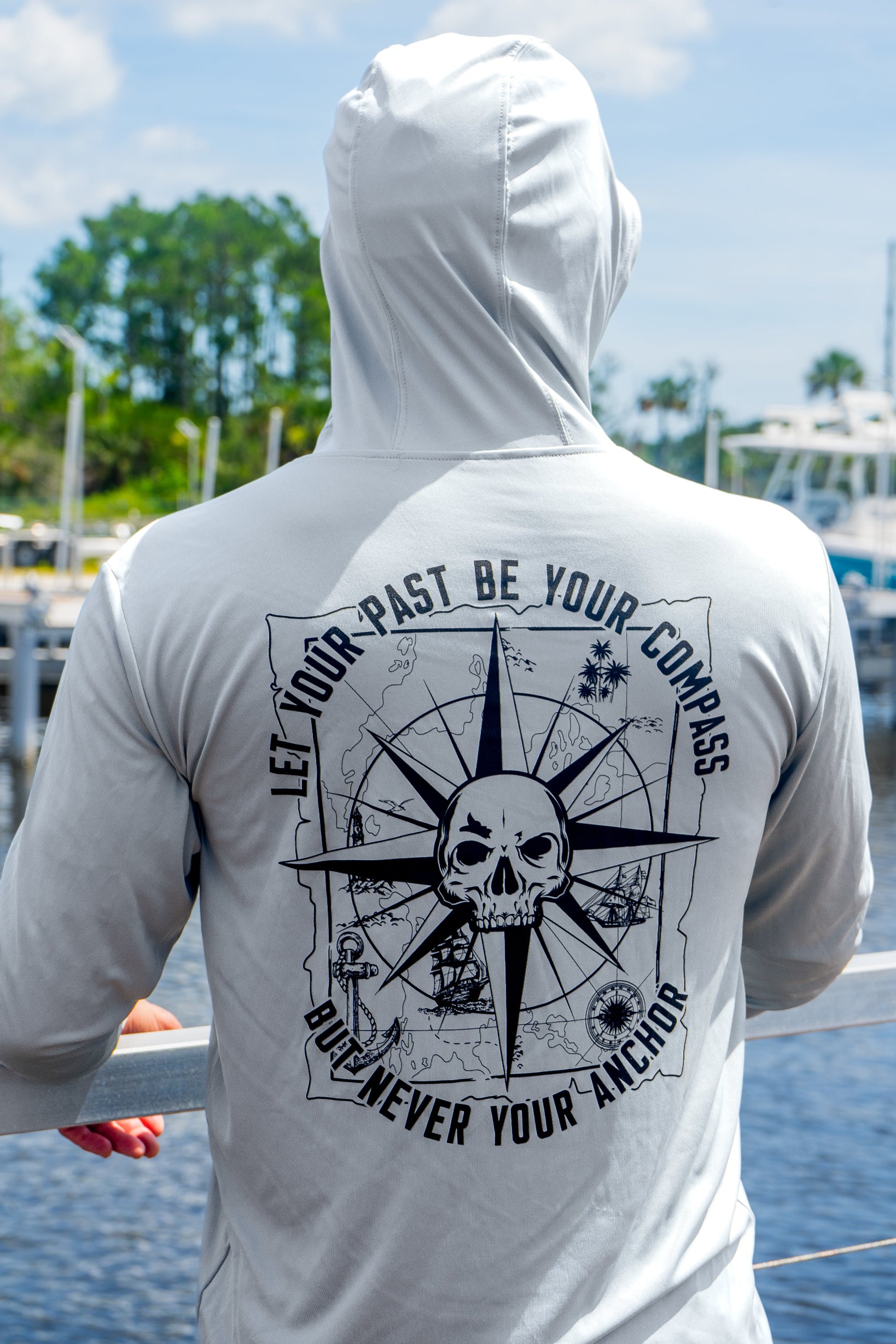 Person wearing a gray hoodie with a skull and compass design on the back, standing by a waterfront.
