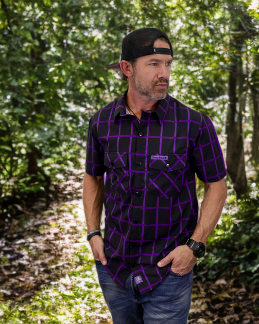Man wearing a black and purple checkered shirt with a brand logo, standing in a forest.