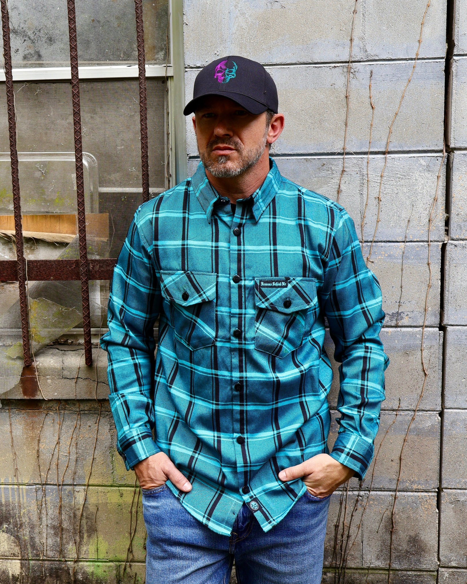 Man wearing a blue plaid shirt and cap standing against a textured wall.
