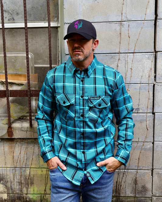 Man wearing a blue plaid shirt and cap standing against a textured wall.
