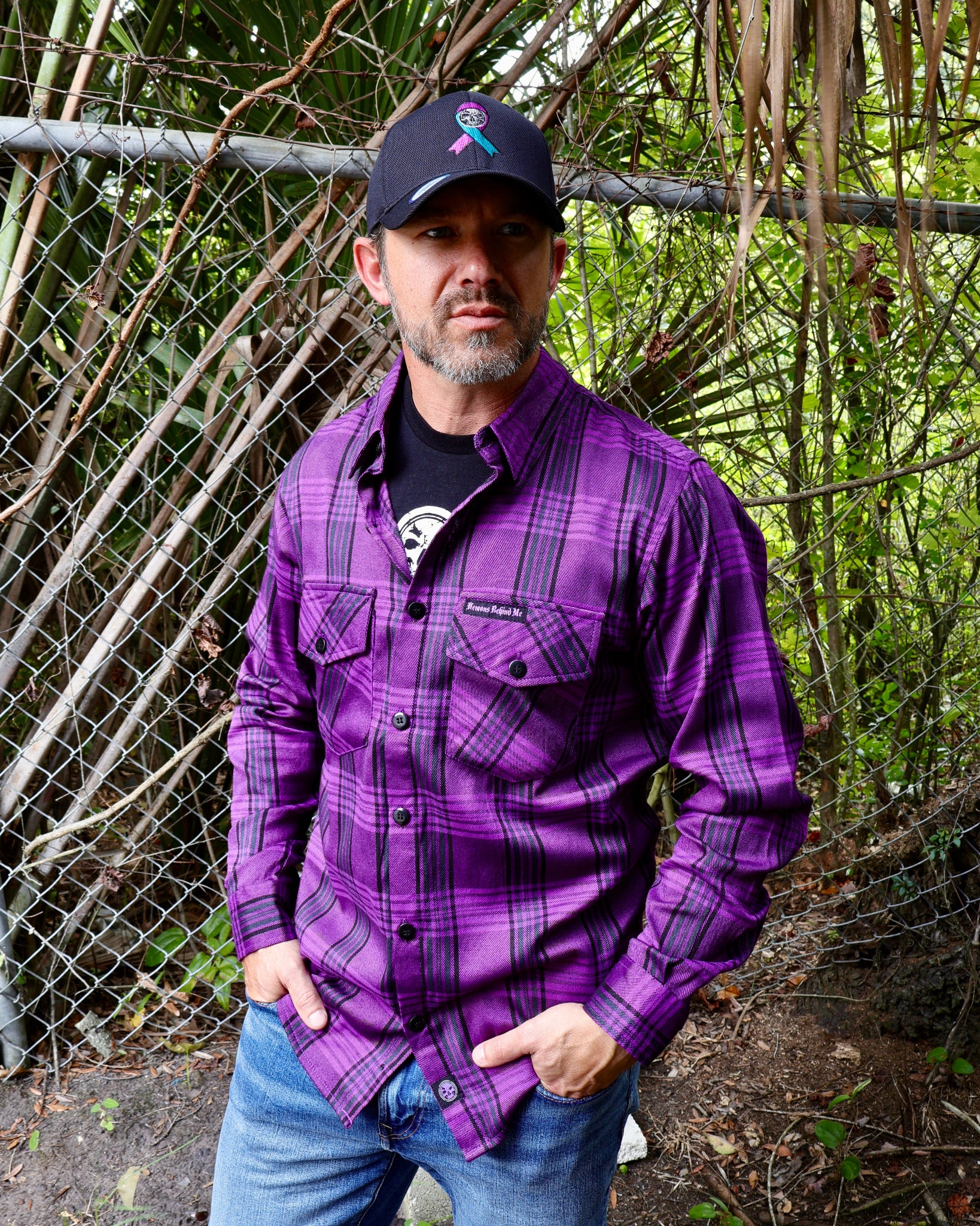 Man wearing a purple plaid shirt and cap with a logo, standing in front of a chain-link fence and trees.