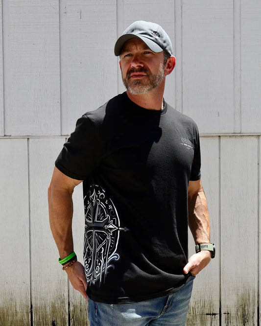 Man wearing a black t-shirt with a white design, standing against a light gray wooden wall.